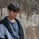 A boy with dark hair leans against a textured wall, looking down thoughtfully.