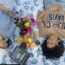 An upside-down view of two smiling female Aggies laying on the grass on an Egghead-designed blanket separated by fruit, flowers and several Egghead branded items including a jigsaw puzzle, cutting board and pen
