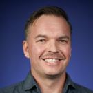 Smiling man with short hair wearing a dark blue button-up shirt, set against a purple background.
