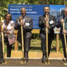Five people in business attire holding gold shovels at an outdoor groundbreaking