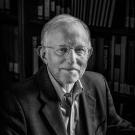 Black and white portrait of an older man with glasses. He wears a button down shirt and jacket, and smiles slightly as he looks into the camera.