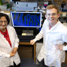 Two smiling scientists in white lab coats by a monitor showing a blue-stained microscope image