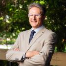 Smiling man in beige suit and glasses, arms crossed, outdoor leafy background
