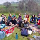 Diverse group of about 20 people picnicking on blankets on a green hillside under gray sky.