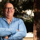 Smiling man with glasses stands confidently against a tree, arms crossed.