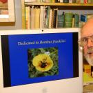 An older man with thinning hair and glasses poses next to a computer screen. On screen is a photo of a bee in the middle of a flower with labeliing "Dedicated to Bombus franklini"