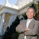 Smiling man in gray blazer, arms crossed, in front of white-columned government building