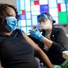 A healthcare worker in protective gear administers a COVID-19 vaccine to a masked individual wearing a black shirt, with a stained-glass window in the background.