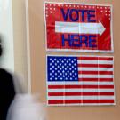 Blurred person walks past red 'VOTE HERE' sign and U.S. flag at polling place