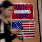 Blurred voter walking past a 'VOTE HERE' sign and American flag in polling place