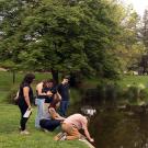 Group of young people at a park pond, some crouching at the water's edge