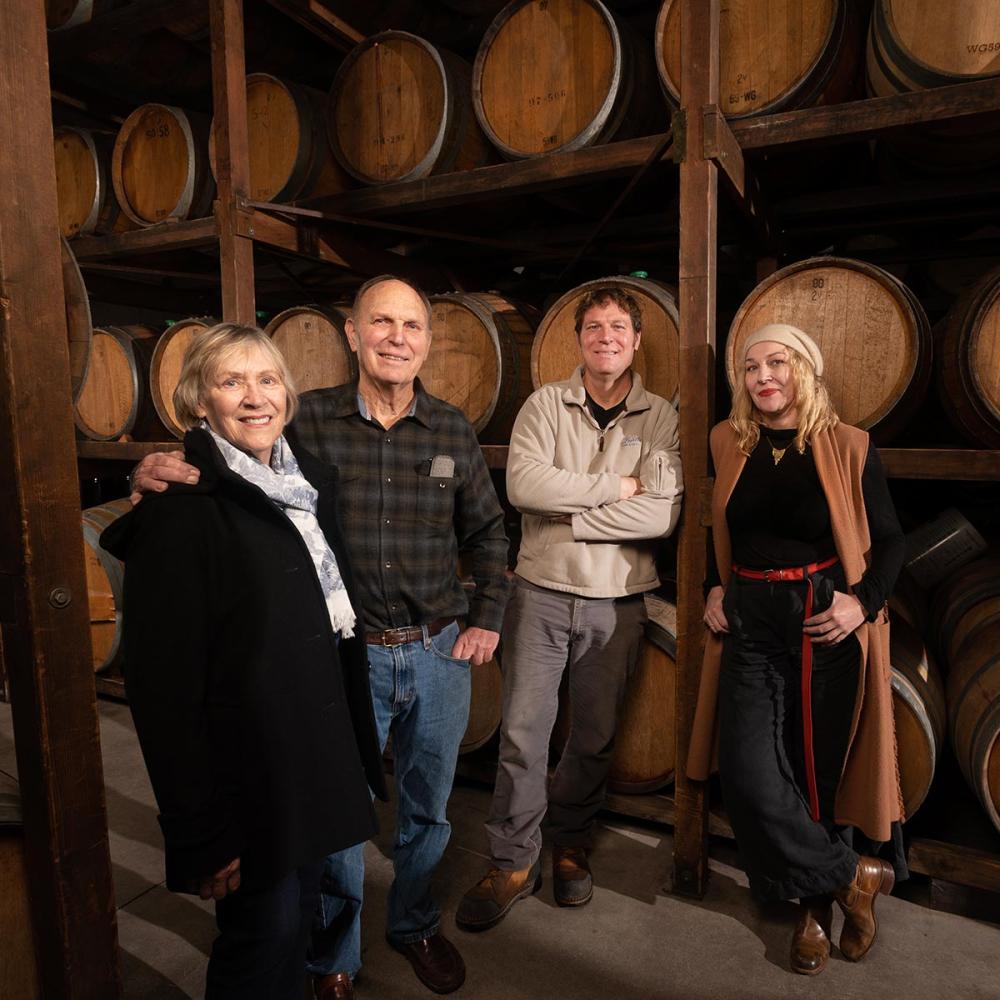 Portrait of four people standing in a large wine cellar, with wine barrels filling the background.